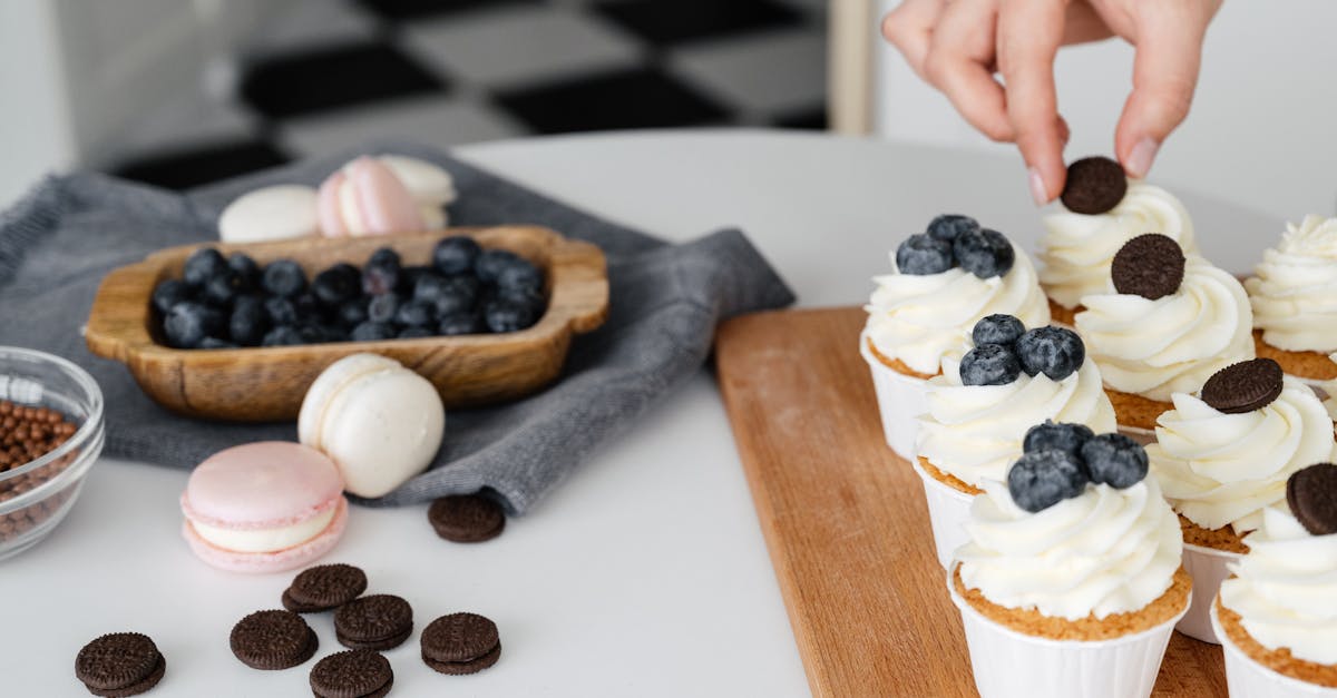 a close up of a hand decorating cupcakes with blueberries and mini cookies in a kitchen setting 1