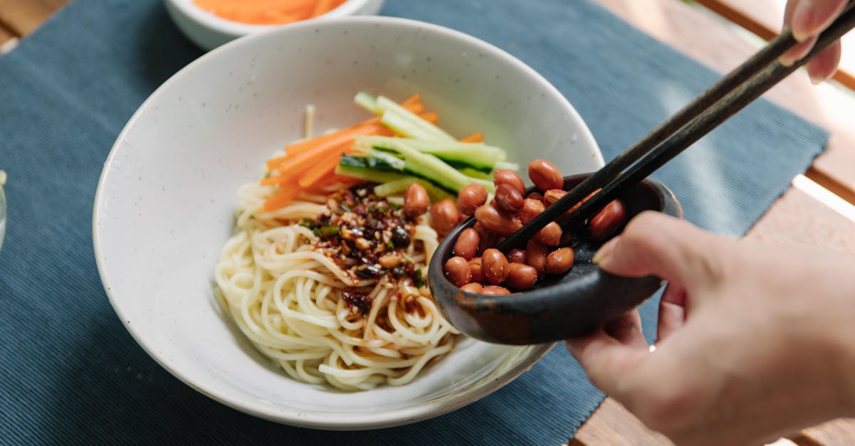 a person adding peanuts to a noodle bowl with vegetables using chopsticks