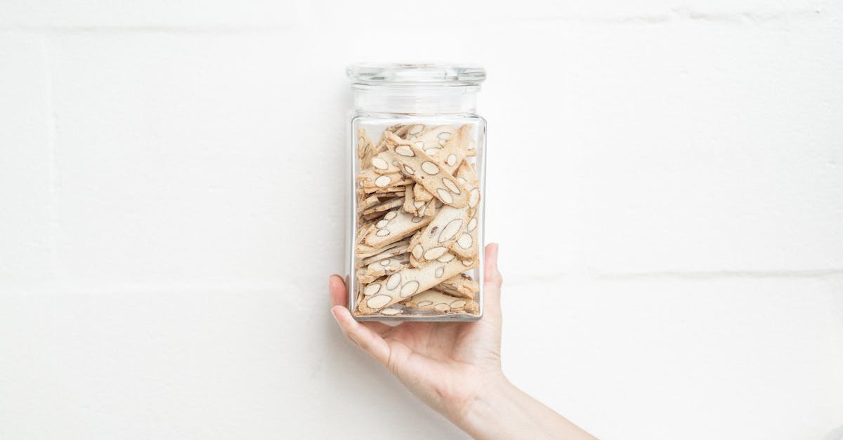 close up of a hand holding a jar filled with almond biscotti against a minimalist white background