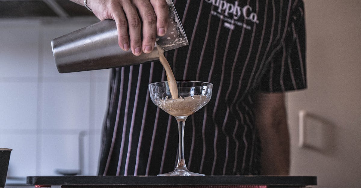 crop anonymous barkeeper pouring tasty alcoholic drink from metal shaker into glass on table in cafe