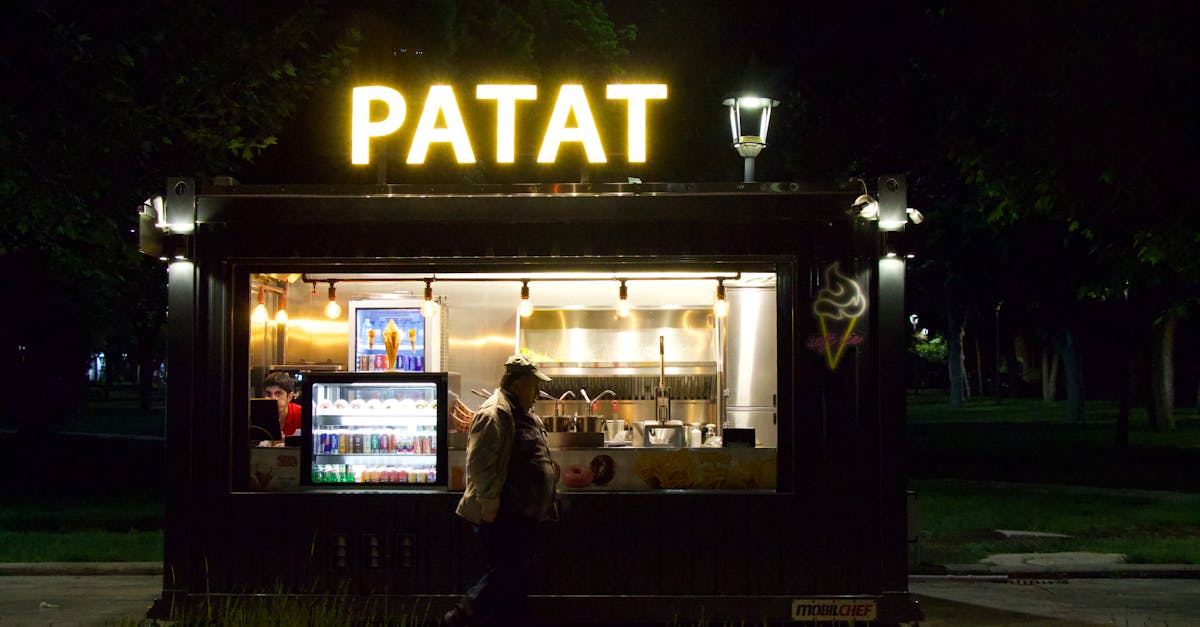 night scene of an illuminated food stall in an urban park with a passerby offering snacks and bever