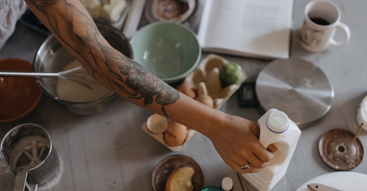 tattooed person pouring milk in a rustic kitchen setting with variety of ingredients on table
